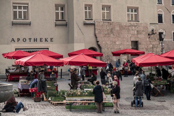 Markt auf gepflastertem Platz mit mehreren roten Sonnenschirmen &uuml;ber Verkaufsst&auml;nden, Menschen, Holzb&auml;nken und Kisten, im Hintergrund eine Geb&auml;udezeile mit dem Schriftzug "APOTHEKE"