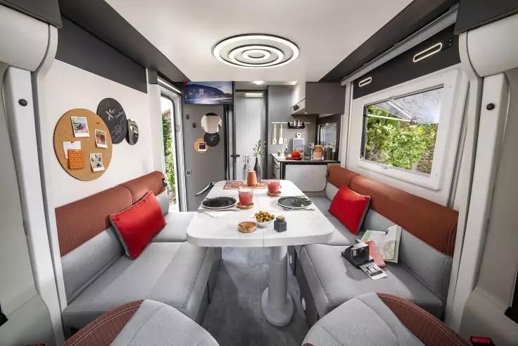 Gegen&uuml;berliegende Sitzb&auml;nke mit Tisch in der Mitte, auf dem Teller, Tassen und eine Karaffe; rote Kissen, Fenster mit gr&uuml;nem Blick, K&uuml;chenzeile mit Schr&auml;nken und Sp&uuml;le, runde Deckenleuchte