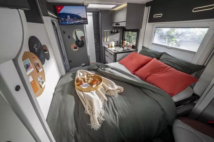 Innenraum mit Bett: dunkelgraue Bettdecke, zwei rote Kissen, wei&szlig;er &Uuml;berwurf; rundes Tablett mit Geb&auml;ck und Getr&auml;nk auf dem Bett; seitliche K&uuml;chenzeile mit Sp&uuml;le und Utensilien, wandmontierter Fernseher, Fenster mit Blick auf B&auml;ume, geschlossene T&uuml;r