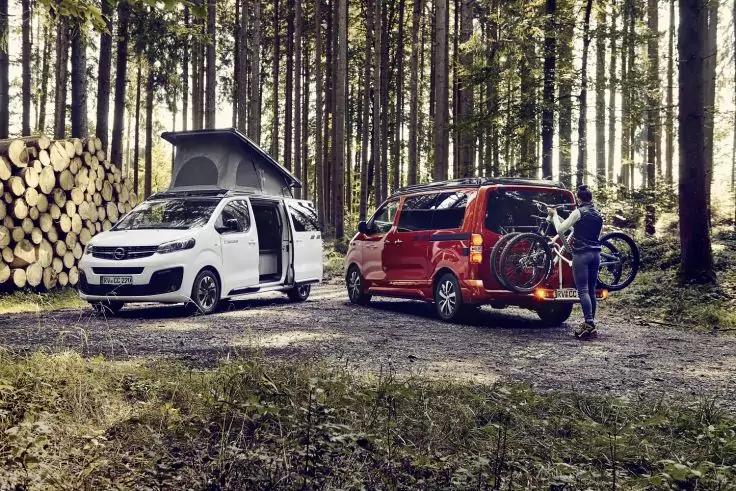 Wei&szlig;es Fahrzeug mit aufgeklapptem Dach, rotes Fahrzeug mit Fahrrad an der Heckklappe, eine Person neben dem Fahrrad, Holzstapel links, Nadelwald im Hintergrund