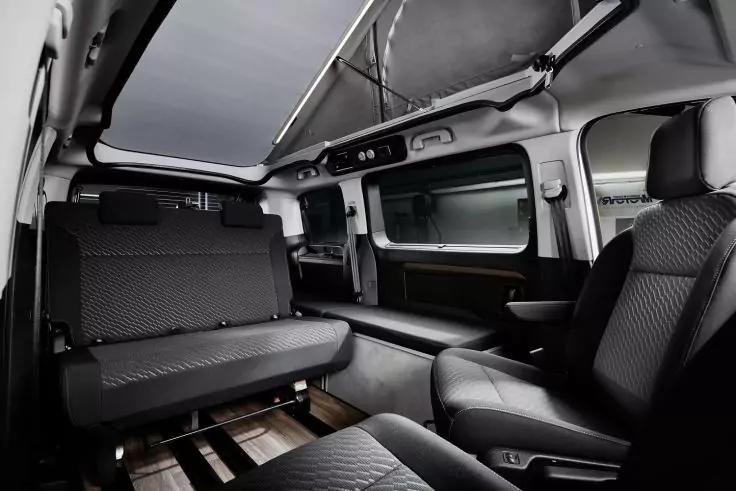 Innenraum eines Fahrzeugs mit grauen gesteppten Sitzen, einer R&uuml;ckbank, einem drehbaren Einzelsitz rechts, gro&szlig;en Seitenfenstern und einem Glasdach