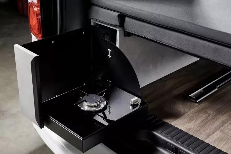 Ausgezogenes schwarzes Metallfach am Heck eines Fahrzeugs mit einem einzelnen Gasbrenner, einem Drehregler, seitlichen Schutzw&auml;nden und Holzoptik-Boden