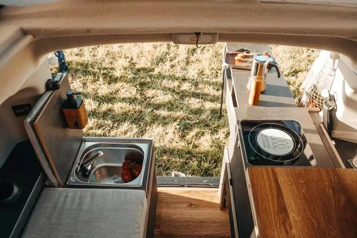 Innenraum eines Fahrzeugs mit offener Heckklappe, kleine K&uuml;chenzeile mit Sp&uuml;le, Herd, Holzarbeitsfl&auml;che und Blick auf eine Wiese