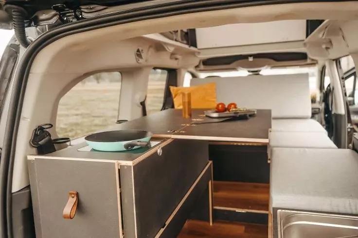 Innenraum eines Fahrzeugs mit offener Heckklappe, eingebauten Holzboxen und Arbeitsfl&auml;che, Bratpfanne, Teller mit Tomaten, gelbes Tuch, Polsterfl&auml;che