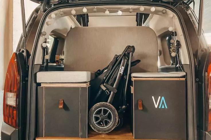 Offener Kofferraum eines Fahrzeugs mit zwei schwarzen Aufbewahrungsboxen, einem gefalteten Kinderwagen in der Mitte und einer beigefarbenen Matratze im Inneren