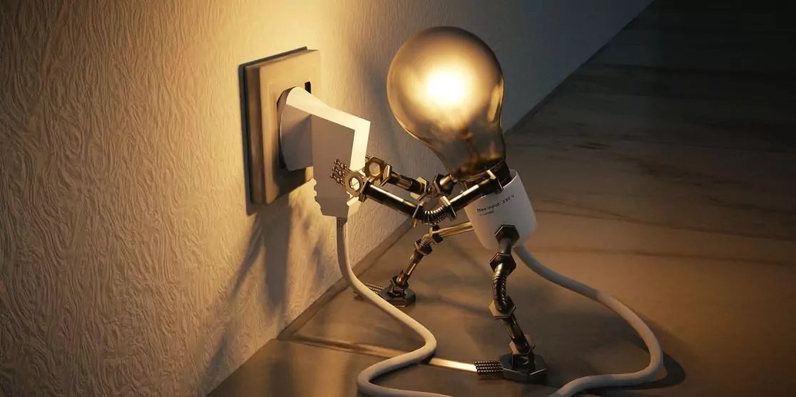 Gl&uuml;hbirne mit Metallgelenken und wei&szlig;em Kabel h&auml;lt einen Stecker, der in eine Wandsteckdose gesteckt wird; warmes Licht an Wand und Holzboden