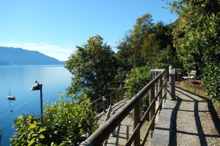 Uferweg mit Gel&auml;nder neben See, B&auml;ume, Laterne, Segelboot auf dem Wasser, Berge im Hintergrund, blauer Himmel