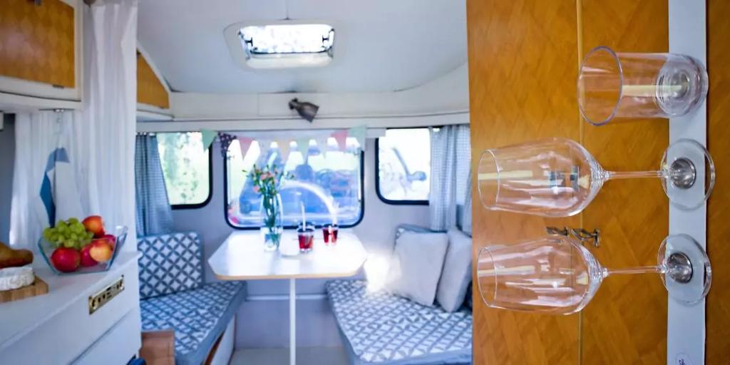 Innenraum mit Sitzecke: Tisch, gepolsterte Sitzb&auml;nke mit gemusterten Kissen, Fenster, Oberlicht, Vorh&auml;nge, Obstschale auf einer Arbeitsfl&auml;che und h&auml;ngende Weingl&auml;ser an einer Holzplatte