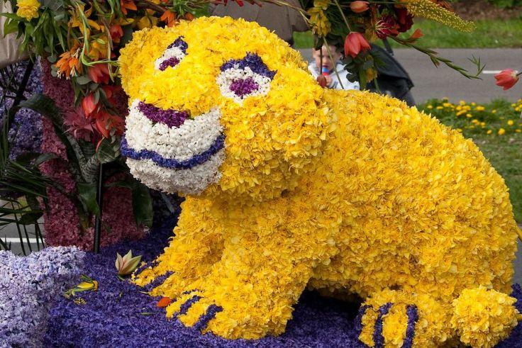 Skulptur in Tierform aus gelben Blumen, mit wei&szlig;en, lila und blauen Gesichtspartien, vier Beinen, auf einem lila-blauen Blumenpodest; im Hintergrund weitere Blumen, Gras und Gehweg