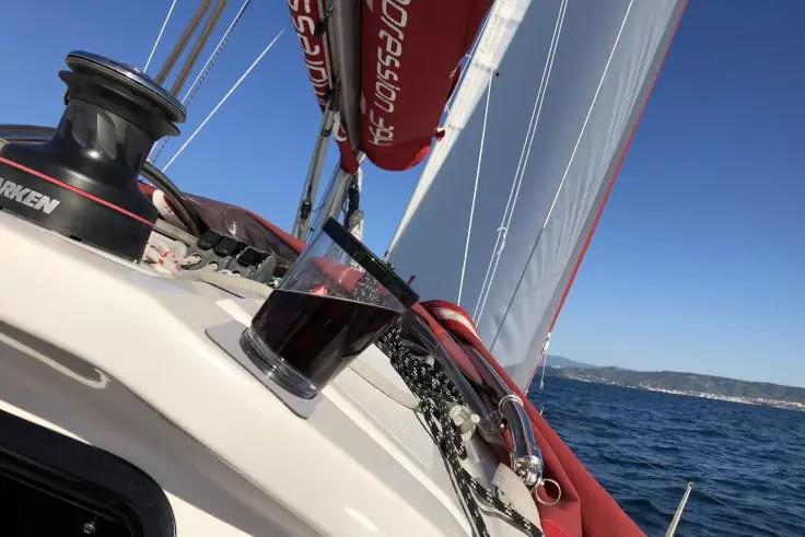 Deck eines Segelbootes mit einem Plastbecher mit dunkler Fl&uuml;ssigkeit auf der Reling, roten und wei&szlig;en Segeln, Taue und blaues Meer am Horizont