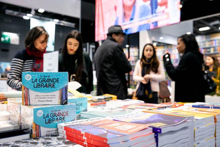 Tisch im Vordergrund mit gestapelten B&uuml;chern, einige Cover zeigen den Titel "La Grande Libraire"; mehrere Personen stehen im Hintergrund, Regale, ein gro&szlig;er Bildschirm und Innenraum sichtbar