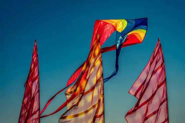 Bunter Drachen mit langen B&auml;ndern und drei rot-gestreiften Fahnen vor blauem Himmel