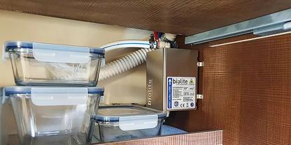 Innen eines Schranks mit drei transparenten, gestapelten Kunststoffbeh&auml;ltern links und rechts eine montierte metallene Box mit Etikett, Kabeln und einem wei&szlig;en, gewellten Schlauch; Holzr&uuml;ckwand sichtbar