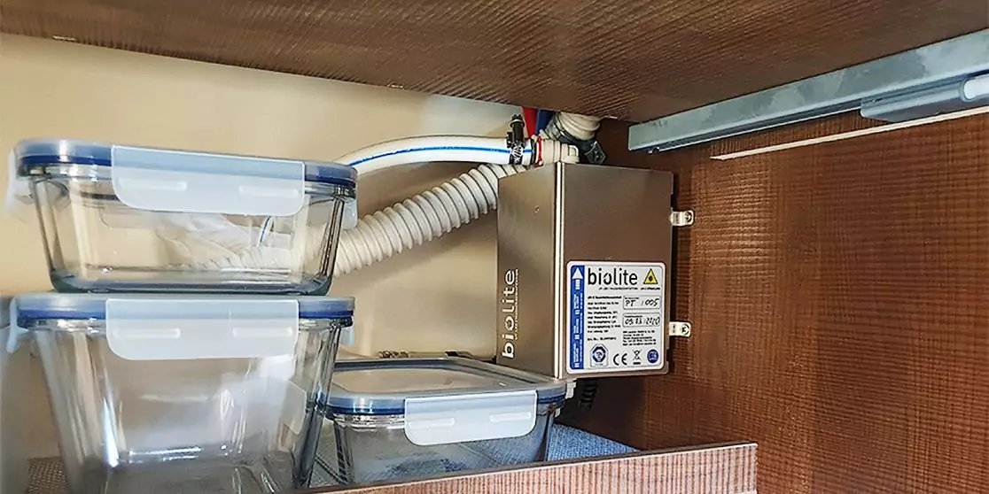 Innen eines Schranks mit drei transparenten, gestapelten Kunststoffbeh&auml;ltern links und rechts eine montierte metallene Box mit Etikett, Kabeln und einem wei&szlig;en, gewellten Schlauch; Holzr&uuml;ckwand sichtbar