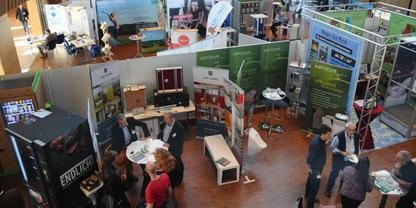 Messest&auml;nde mit Rollups, Tischen und Displays, mehrere Personen in der Halle, Holzfu&szlig;boden