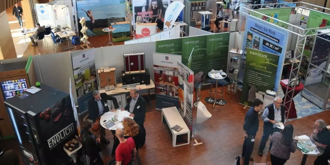 Messest&auml;nde mit Rollups, Tischen und Displays, mehrere Personen in der Halle, Holzfu&szlig;boden