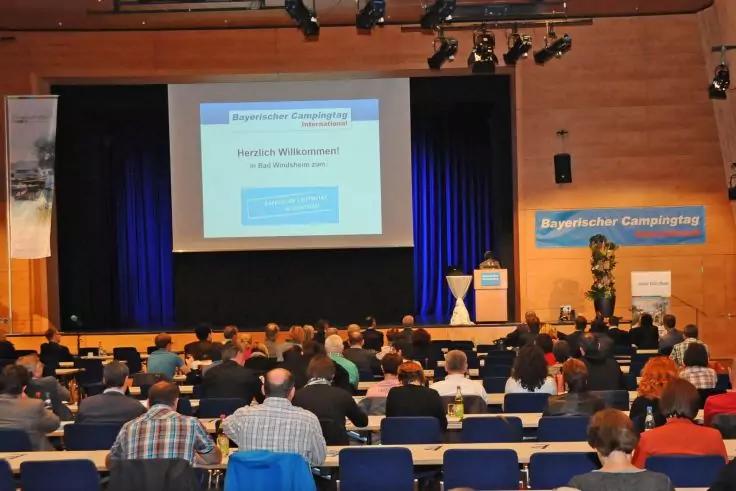 Konferenzsaal mit Reihen sitzender Personen, Blick zur B&uuml;hne; Projektionsleinwand &uuml;ber der B&uuml;hne, Person am Rednerpult, blaue Vorh&auml;nge, Holzverkleidung, Banner rechts