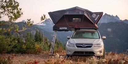 Wei&szlig;es SUV mit aufgeklapptem Dachzelt und Leiter auf felsiger Wiese, Nadelb&auml;ume links, Berggipfel mit Schneeflecken im Hintergrund, orange-rosa Himmel