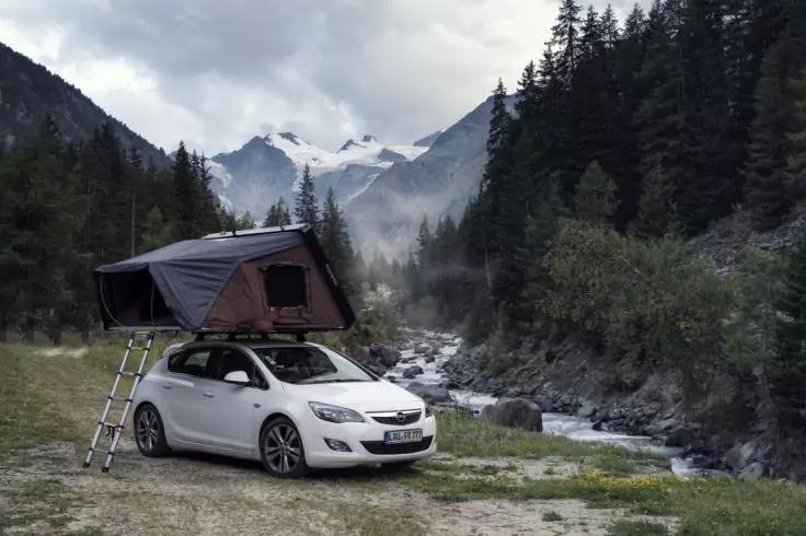 Wei&szlig;es Auto mit aufgeschlagenem Dachzelt und Leiter neben einem Bach in einem bergigen, bewaldeten Tal, im Hintergrund schneebedeckte Gipfel und Nebel