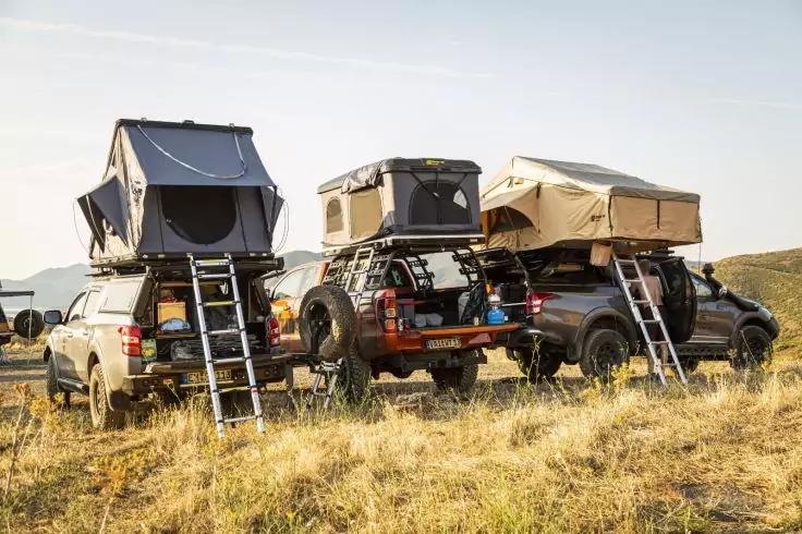 Drei Pickup-Fahrzeuge auf trockenem Grasfeld, jeweils mit ausgeklappten Dachzelten und Leitern; offene Zelten, Dachtr&auml;ger und H&uuml;gel im Hintergrund unter klarem Himmel
