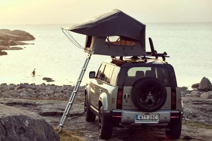 Gel&auml;ndewagen mit aufgeklapptem Dachzelt und ausgezogener Leiter, geparkt auf felsigem Ufer, Meer im Hintergrund, Person am Wasser