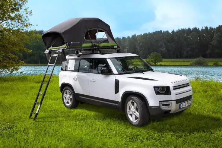 Wei&szlig;er SUV mit ge&ouml;ffnetem Dachzelt und angelehnter Leiter auf gr&uuml;ner Wiese neben einem See, Wald im Hintergrund, klarer Himmel