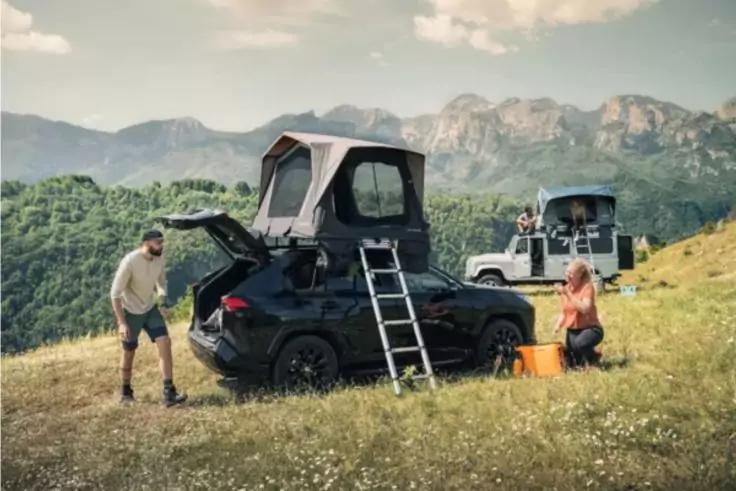 Schwarzes Auto mit aufgeklapptem Dachzelt und Leiter, offene Heckklappe; Personen neben dem Fahrzeug auf einer Wiese, im Hintergrund zweites Auto mit Dachzelt und Berglandschaft