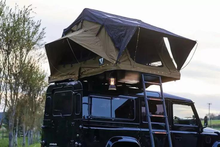 Schwarzes Gel&auml;ndefahrzeug mit aufgeklapptem Dachzelt, ausfahrbarer Leiter, Wiese, B&auml;ume und bew&ouml;lkter Himmel