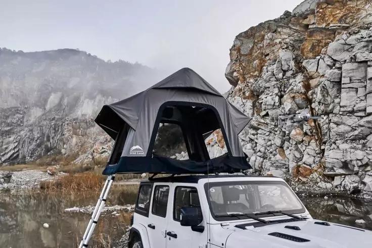 Wei&szlig;es Gel&auml;ndefahrzeug mit ge&ouml;ffnetem Dachzelt und Leiter, neben einer Wasserfl&auml;che an einer felsigen Klippe, Berge und Nebel im Hintergrund