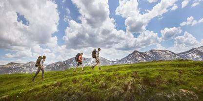 Drei Personen mit Rucks&auml;cken gehen auf einem grasbewachsenen Bergr&uuml;cken, im Hintergrund schneebedeckte Berge und bew&ouml;lkter Himmel