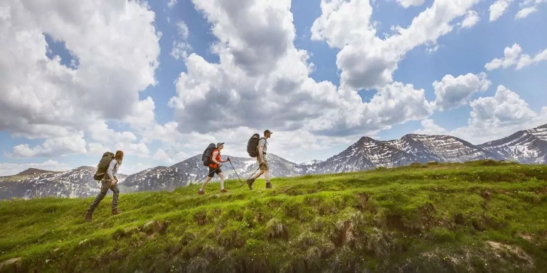 Drei Personen mit Rucks&auml;cken gehen auf einem grasbewachsenen Bergr&uuml;cken, im Hintergrund schneebedeckte Berge und bew&ouml;lkter Himmel