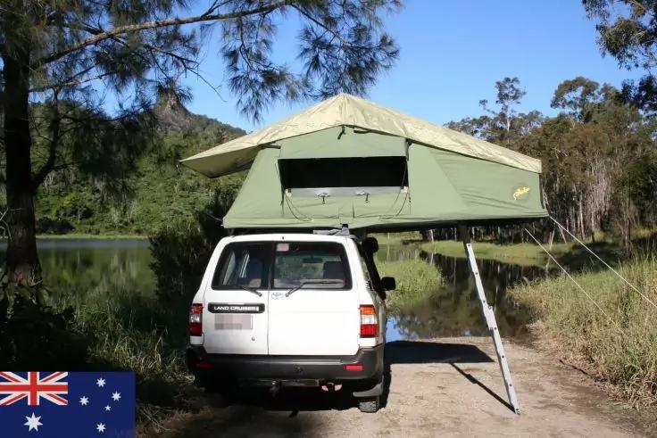 Wei&szlig;er SUV mit ge&ouml;ffnetem gr&uuml;nem Dachzelt und Leiter auf einem Feldweg neben einem See, B&auml;ume im Hintergrund, australische Flagge unten links