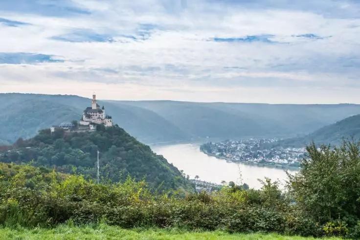 Burg auf bewaldetem H&uuml;gel links, Fluss mit einer Schlinge in der Bildmitte, Ortschaft am rechten Ufer, Wiese und B&uuml;sche im Vordergrund, bew&ouml;lkter Himmel