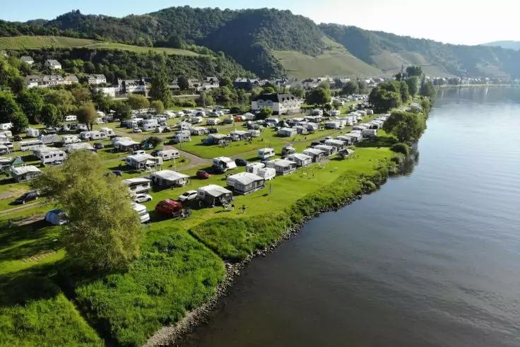 Reihen von Wohnwagen und Wohnmobilen auf einer grasbewachsenen Fl&auml;che am Flussufer, Wasser rechts, H&uuml;gel im Hintergrund