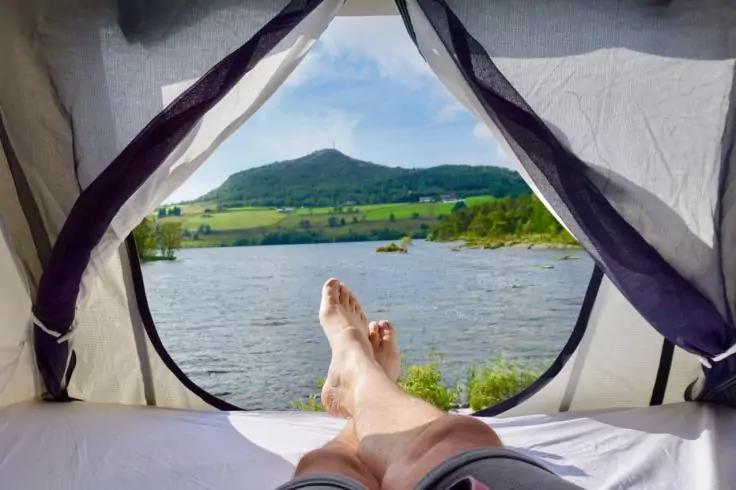 Blick aus Zelt&ouml;ffnung: ausgestreckte Beine und nackte F&uuml;&szlig;e, See, grasiges Ufer, B&auml;ume, H&uuml;gel und Himmel mit Wolken