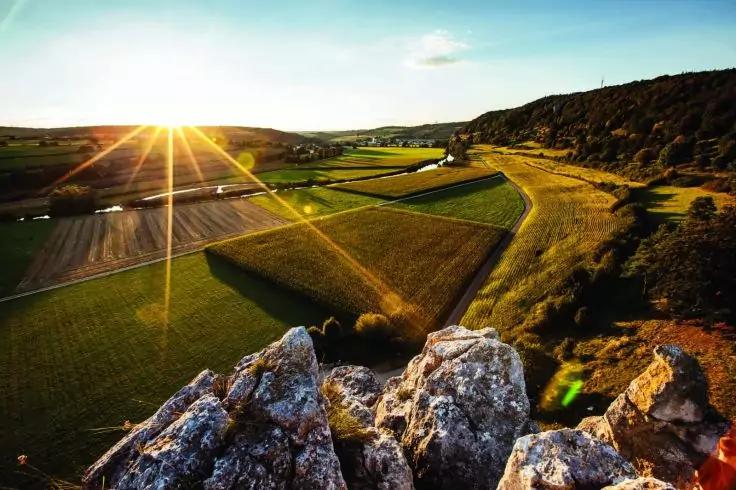 Felsvorsprung im Vordergrund, darunter rechteckige Feldparzellen, gewundene Landstra&szlig;e, H&uuml;gel rechts, niedrige Sonne mit sichtbaren Strahlen am Horizont, vereinzelte Geb&auml;ude