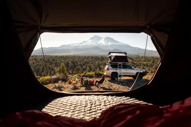 Blick aus dem Inneren eines Zeltes auf ein Campingplatz mit Pickup und Dachzelt, zwei Campingst&uuml;hle und ein kleiner Tisch, Gestr&uuml;pp und Wald, im Hintergrund ein schneebedeckter Berg