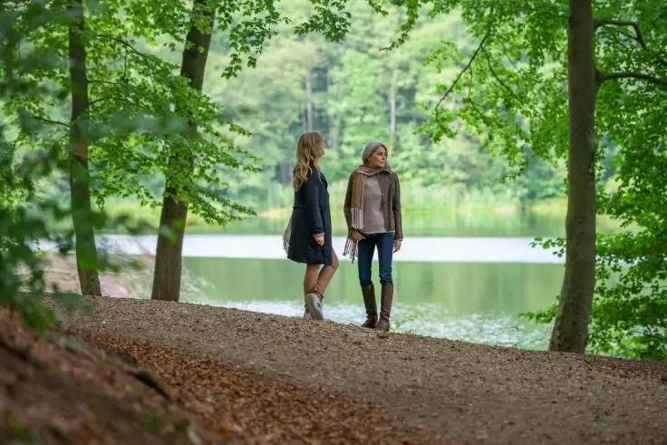 Zwei Frauen stehen auf einem mit Laub bedeckten Waldweg neben einem See, umgeben von gr&uuml;nen B&auml;umen