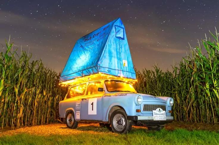 Hellblaues Auto mit aufgeklapptem blauem Dachzelt, beleuchtet von gelben Lichterketten, geparkt auf Gras zwischen hohen Maispflanzen unter einem sternklaren Nachthimmel. Nummer 1 an der T&uuml;r