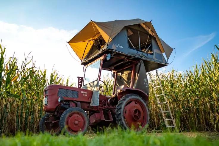Roter Traktor mit aufgesetztem Dachzelt, an dem eine Leiter lehnt, vor hohem Maisfeld und blauem Himmel