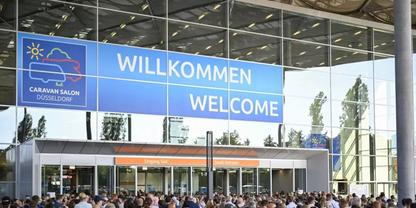 Gro&szlig;e Glasfassade mit blauem Schild 'WILLKOMMEN WELCOME', kleinem Logo, Menschenmenge vor dem Eingang, S&auml;ulen und Spiegelungen im Glas