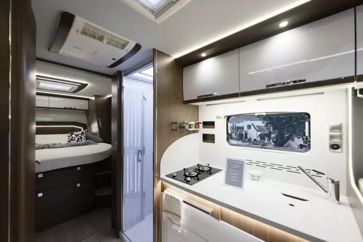 Innenraum mit K&uuml;chenzeile: Sp&uuml;le mit Armatur, zweiflammiger Herd, Arbeitsfl&auml;che, Fenster, H&auml;ngeschr&auml;nke und Beleuchtung; im Hintergrund Durchgang zur Schlafnische mit Bett und Kissen