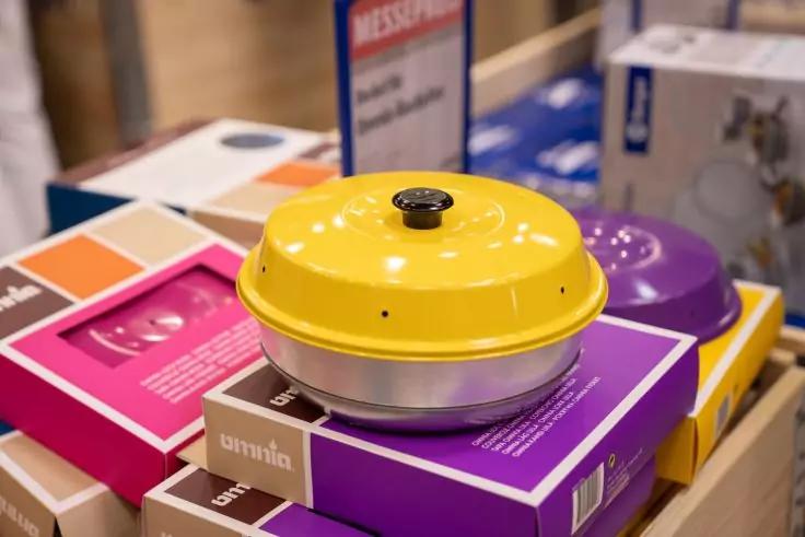 Gelbe runde Pfanne mit Deckel auf gestapelten bunten Produktverpackungen; im Hintergrund eine violette Pfanne und Regale