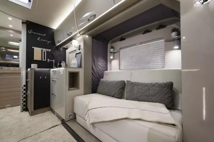 Innenraum mit hellem Sofa, zwei grauen Kissen, gefalteter Decke, Fenster mit Jalousie, Oberschr&auml;nken mit Beleuchtung und kompakter K&uuml;chenzeile mit Sp&uuml;le und Schr&auml;nken