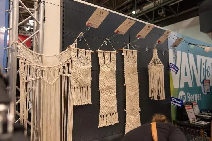 F&uuml;nf beige Makramee-Wandbeh&auml;nge unterschiedlicher L&auml;nge an einer Metallstange vor schwarzer Trennwand, unten rechts der R&uuml;cken einer gebeugten Person, im Hintergrund ein Messestand mit bunten Schildern