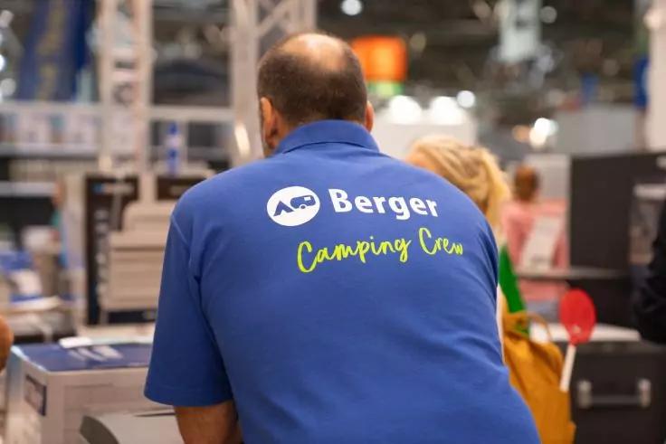 Person von hinten mit blauem Poloshirt, auf dem hinten 'Berger Camping Crew' und ein rundes Logo zu sehen sind; im Hintergrund unscharfe St&auml;nde, Ger&auml;te und Personen