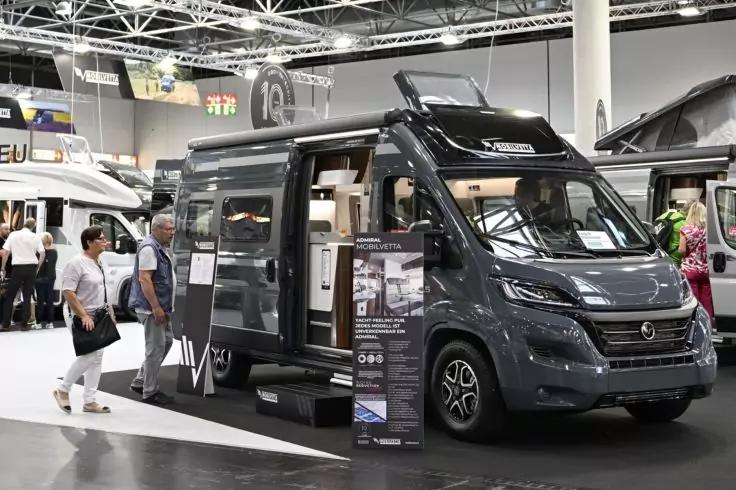 Dunkelgrauer Campervan in Ausstellungshalle, seitliche Schiebet&uuml;r ge&ouml;ffnet, Blick ins Innere, Informationsaufsteller davor, weitere Fahrzeuge und Besucher im Hintergrund