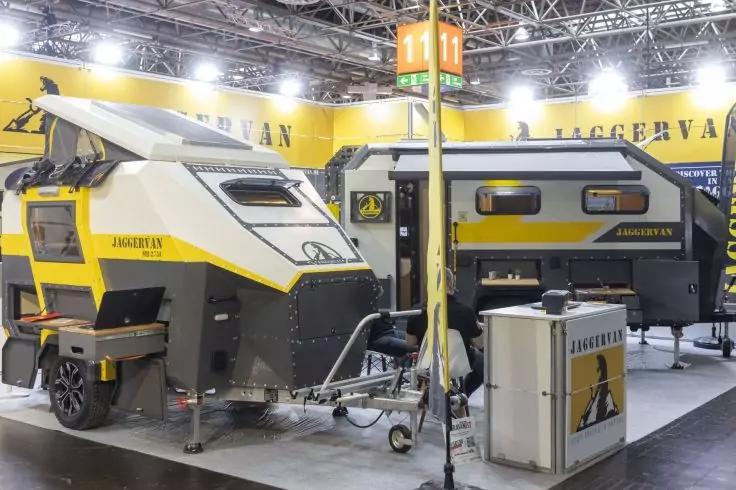 Zwei gelb-graue Camping-Anh&auml;nger in einem hell beleuchteten Messestand, Informationspult und Deckenbeleuchtung sichtbar