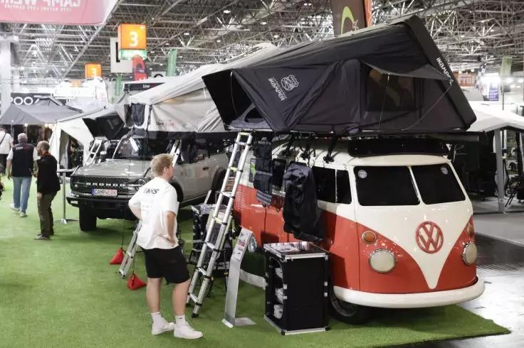Roter und wei&szlig;er Volkswagen-Bus mit aufgeklapptem Dachzelt, Leiter an der Seite, eine Person steht daneben, weitere Fahrzeuge und Ausstellungshalle im Hintergrund