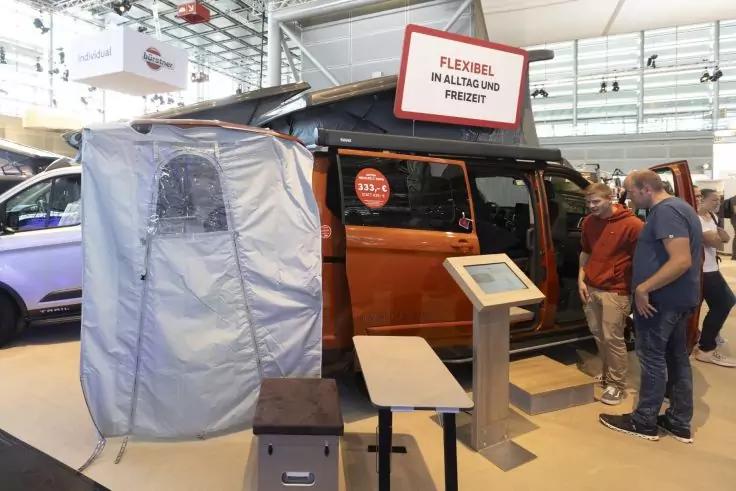 Orangefarbener Van mit ge&ouml;ffnetem Seitendach und grauem Zeltanbau, Schild auf dem Dach mit Text FLEXIBEL IM ALLTAG UND FREIZEIT, Touchscreen-Informationsstand vorne, zwei Personen rechts